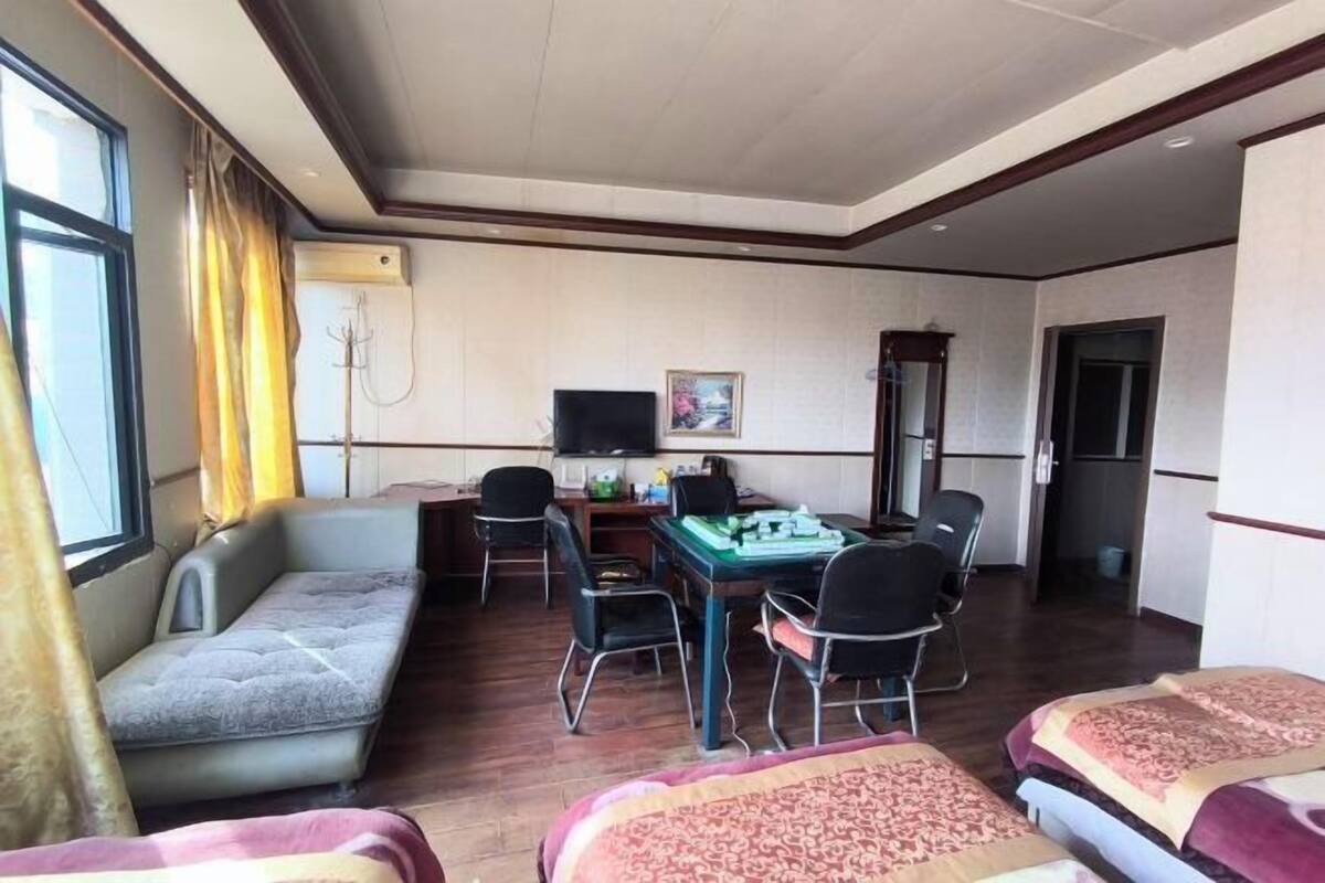 A room with a sofa, two single beds, a desk with chairs, a television, and a window with curtains.