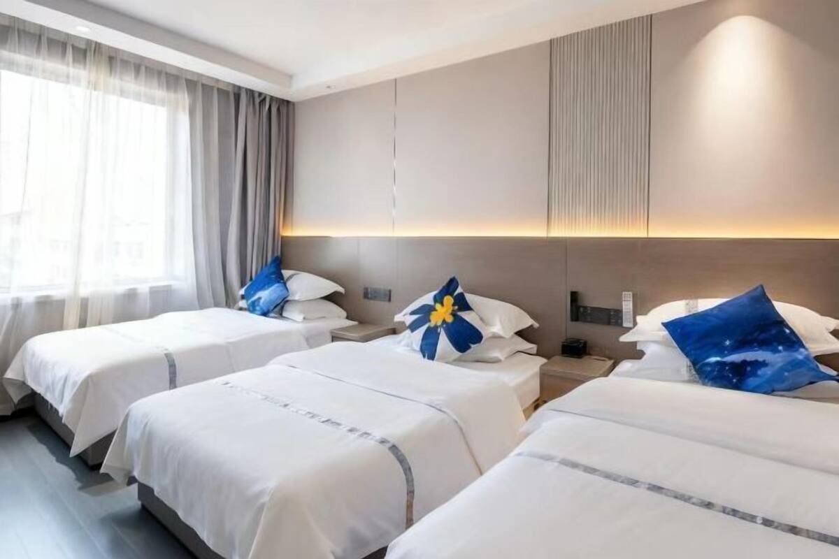 A hotel room with three beds, a large window with curtains, and a wall-mounted light.