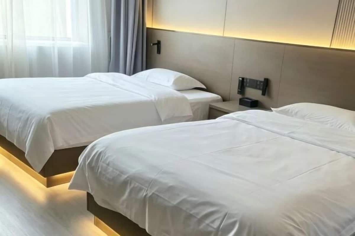 A hotel room with two beds, a bedside table, a wall-mounted control panel, and a window with curtains.