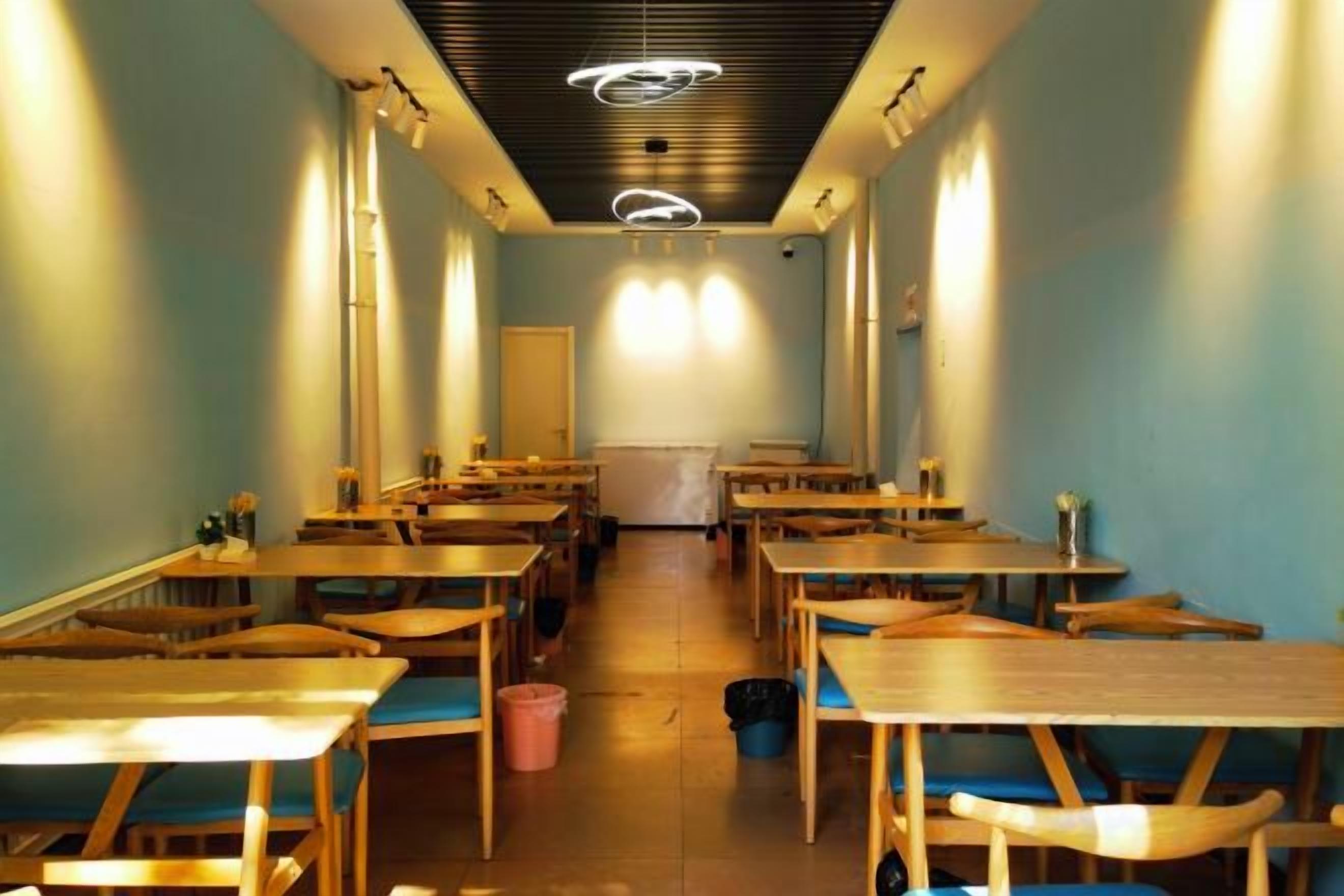 Une longue pièce étroite, avec des rangées de tables et de chaises en bois, une allée centrale et un plafond orné de luminaires circulaires.