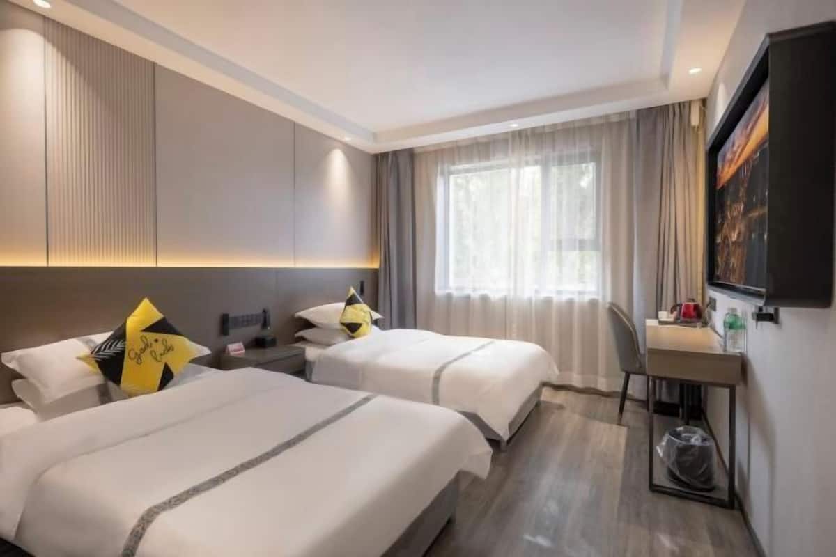 A hotel room with two beds, a desk, a chair, a window with curtains, and a wall-mounted TV.