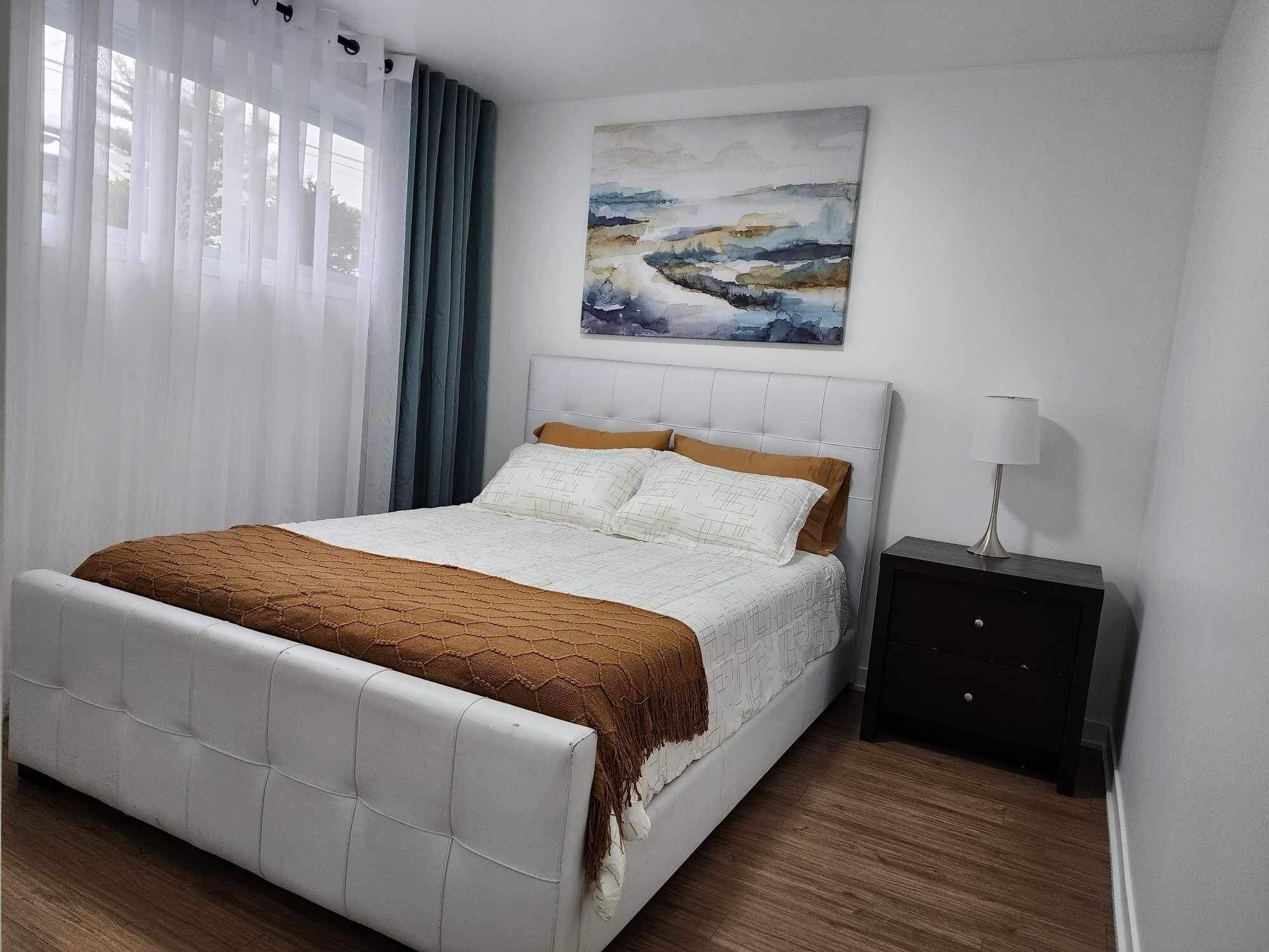 A bedroom with a bed, a nightstand, a painting, and a window with curtains.