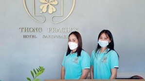 Deux employés de l’hôtel Thong Prachok se tiennent derrière le comptoir d’accueil, devant une enseigne portant le nom de l’hôtel.
