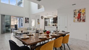 A spacious dining area with a long table set for four, a kitchen in the background, and a large window providing natural light. - Large 5BR Home w Pool, Princess Room, Resort Access (Kissimmee)