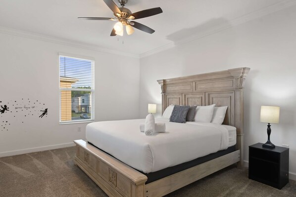 A bedroom with a large bed, a ceiling fan, a nightstand with a lamp, and a window with blinds.