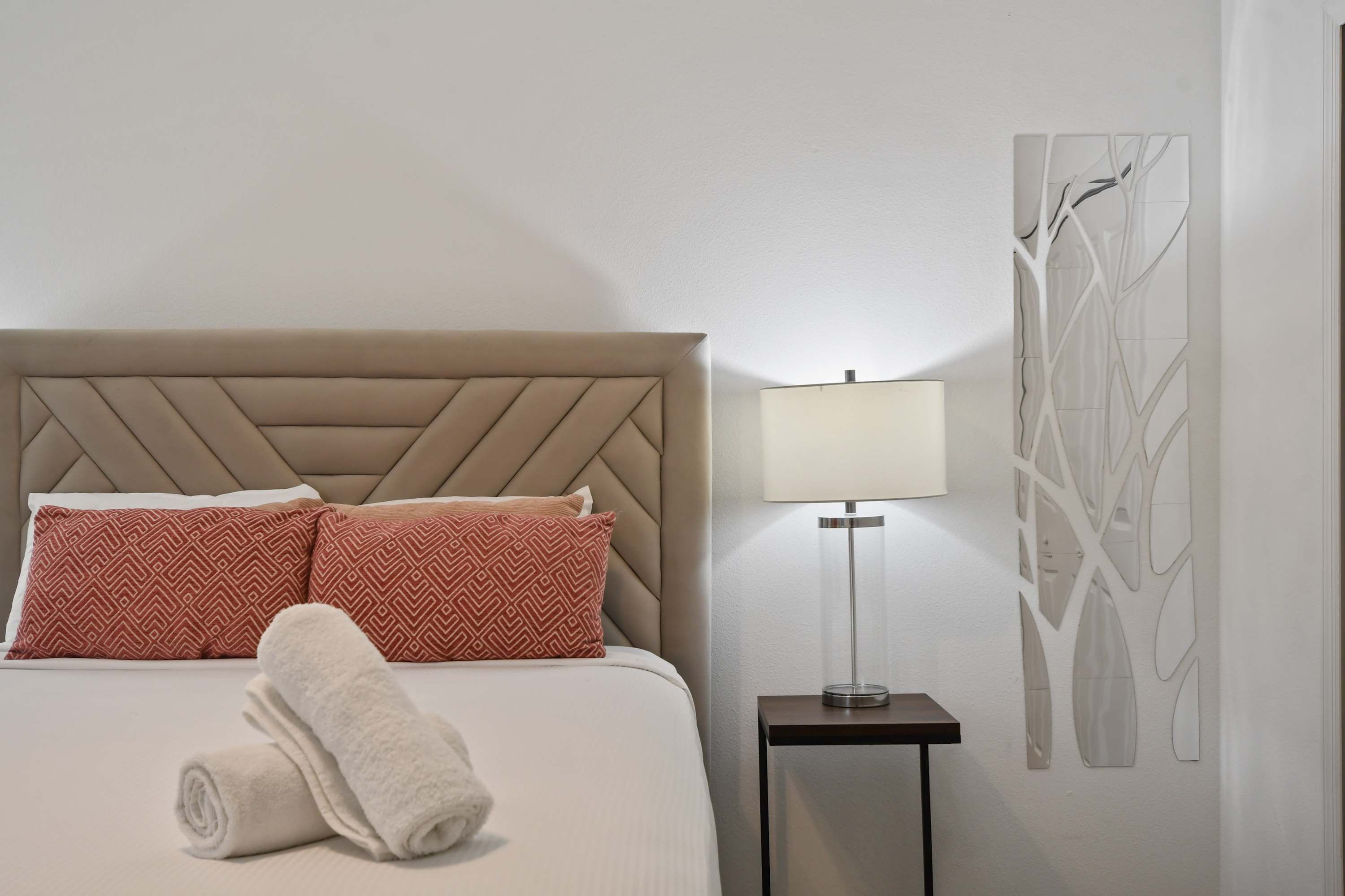 A neatly made bed with patterned pillows, a rolled towel, and a bedside table with a lamp.