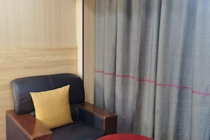 A room with a dark blue leather armchair, a wooden side table, a yellow pillow, and a grey curtain with a red stripe. - Shankee Hotel (Nanchang Chengbihu Park) (Nanchang County)