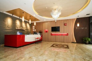 Zona de recepción de un hotel con mostrador rojo, pared de madera, candelabro y pantalla de televisión.