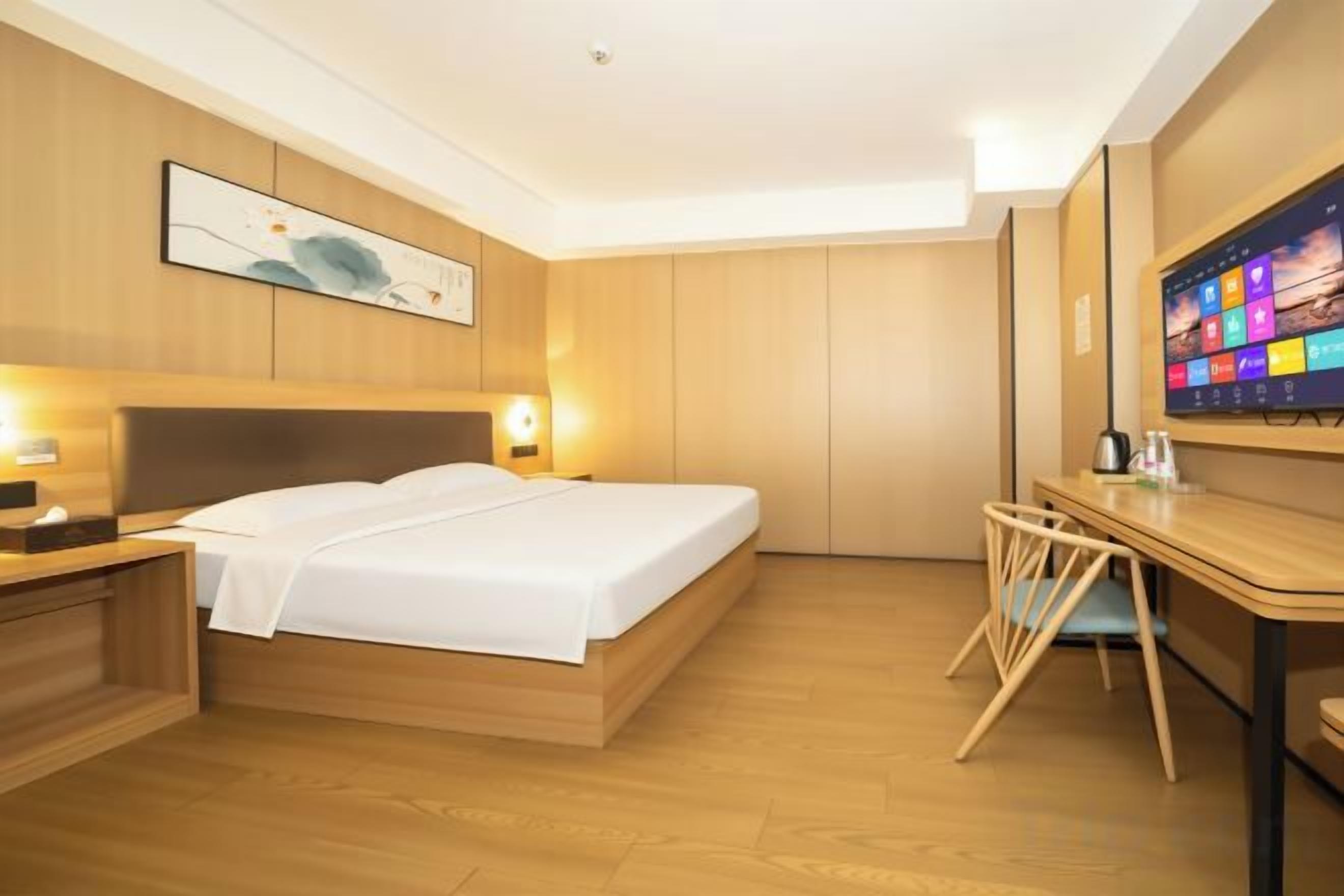 A hotel room with a bed, bedside table, desk, chair, and a large digital screen on the wall.