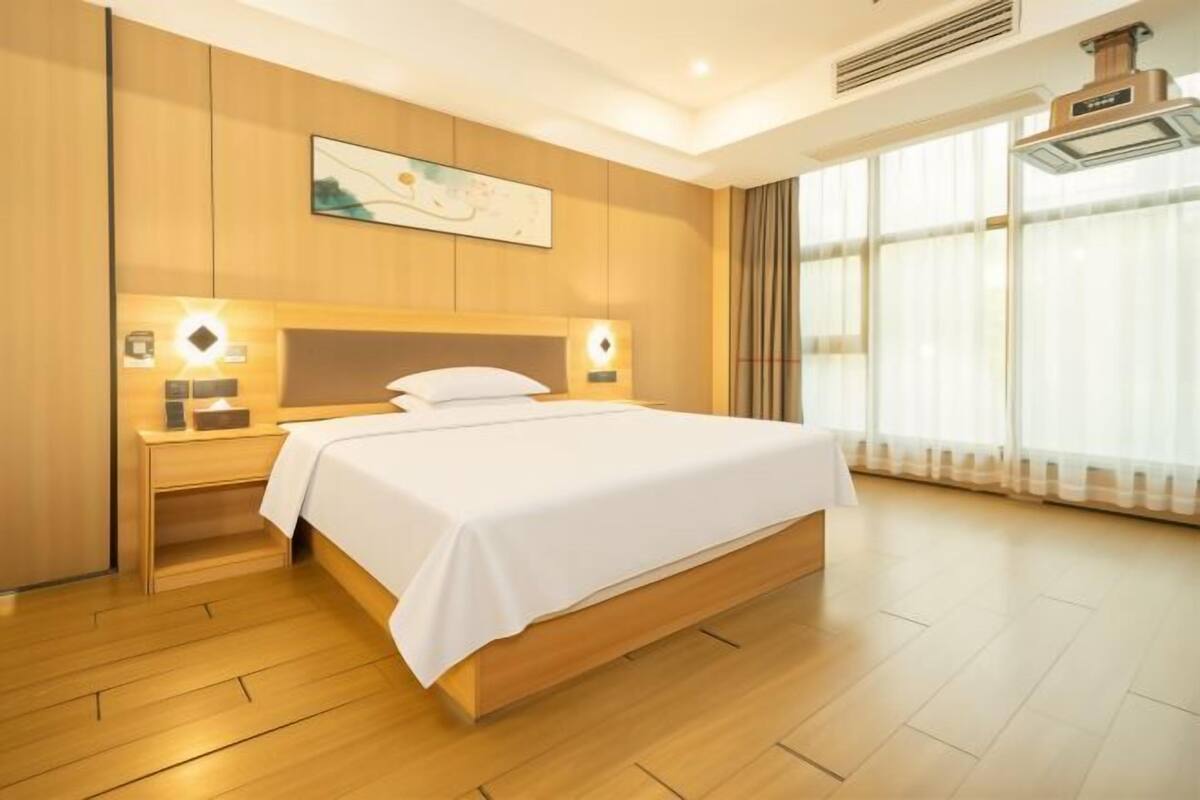 Een hotelkamer met een groot bed, houten lambrisering, een nachtkastje met lamp en een groot raam met gordijnen.