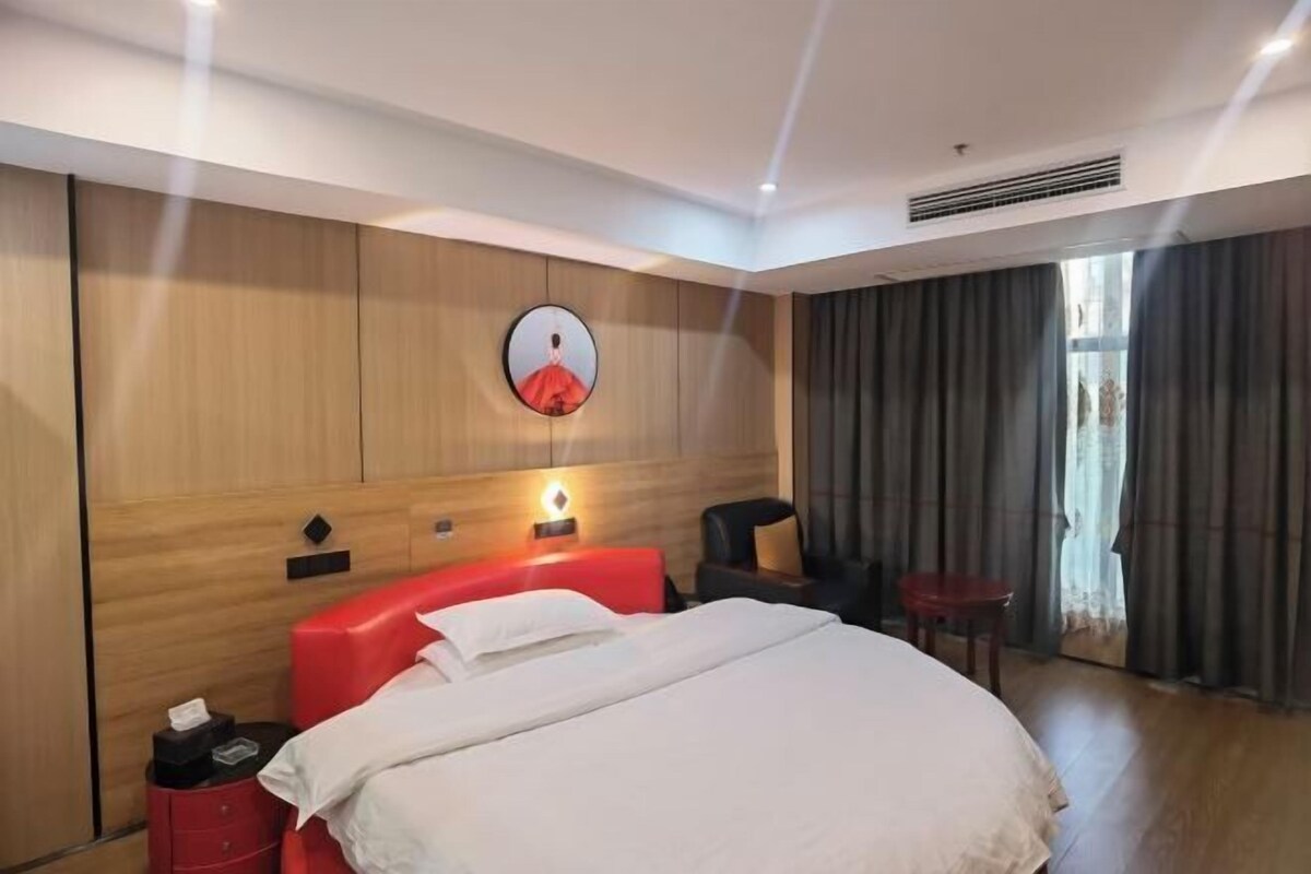 A hotel room with a bed, a red headboard, a wall-mounted TV, a bedside table, and a window with curtains.