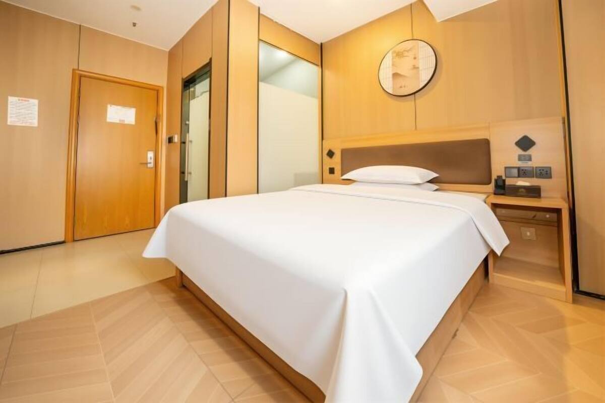 Een hotelkamer met een groot bed, een houten hoofdeinde, een nachtkastje met een lamp en een wandklok.