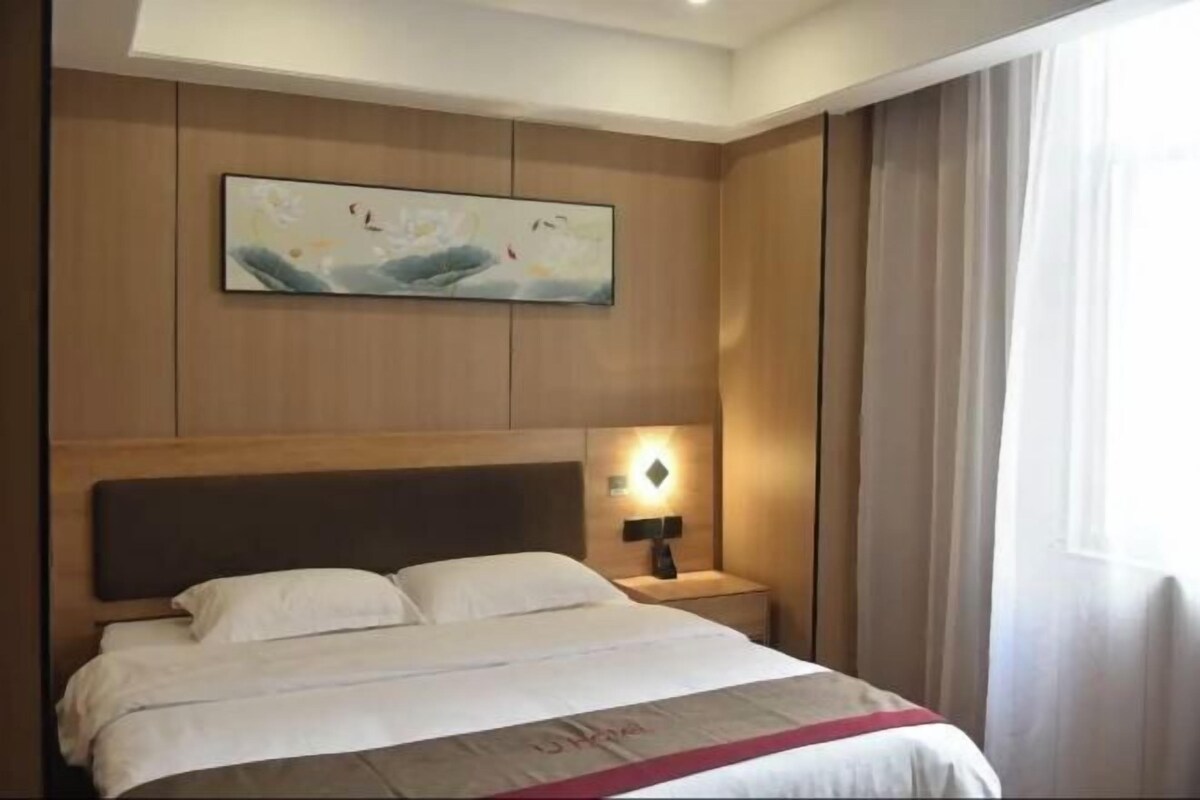 A hotel room with a bed, a nightstand, a wall-mounted lamp, and a painting above the bed.