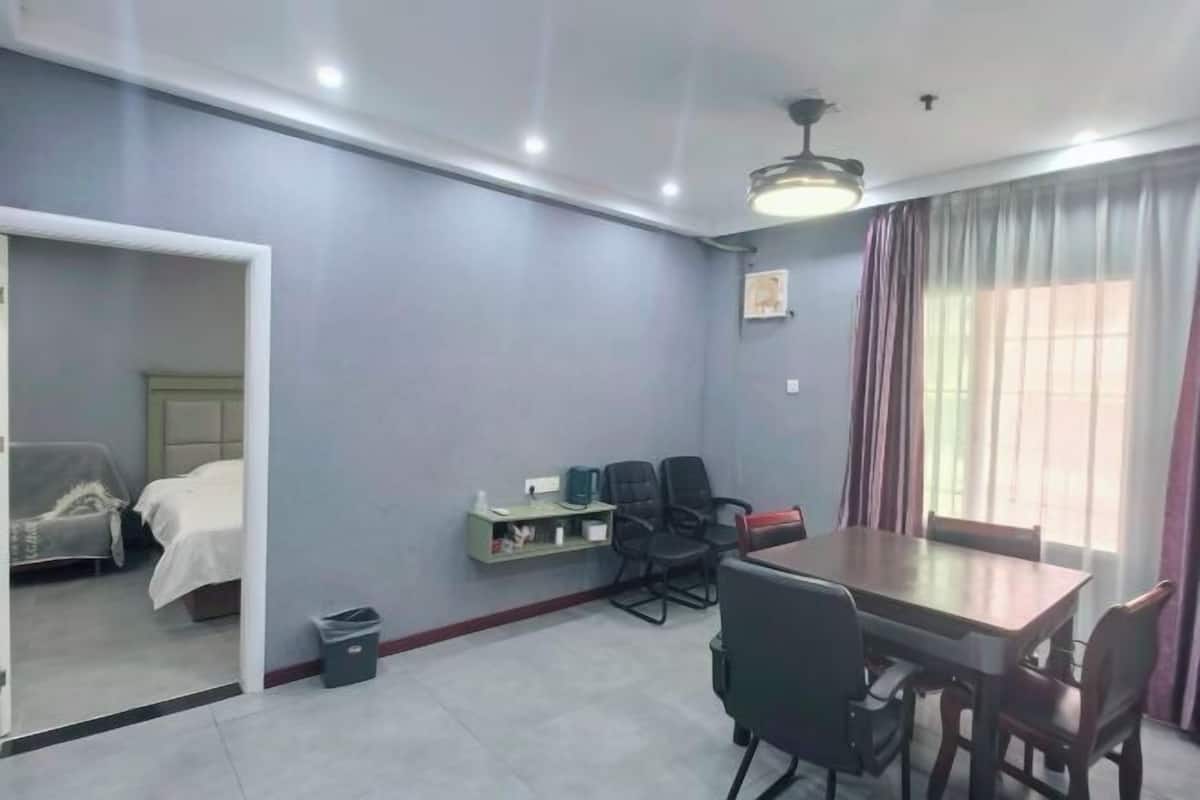 A room with a bed, a desk with chairs, a small shelf, and a window with curtains.