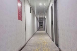 Um corredor comprido e estreito com carpete estampado, portas em ambos os lados e uma placa vermelha na parede esquerda.