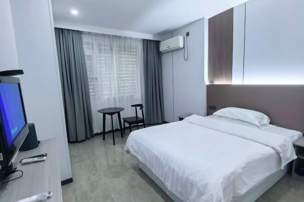 A hotel room with a bed, a TV, a desk with a chair, a small table, and a window with curtains.