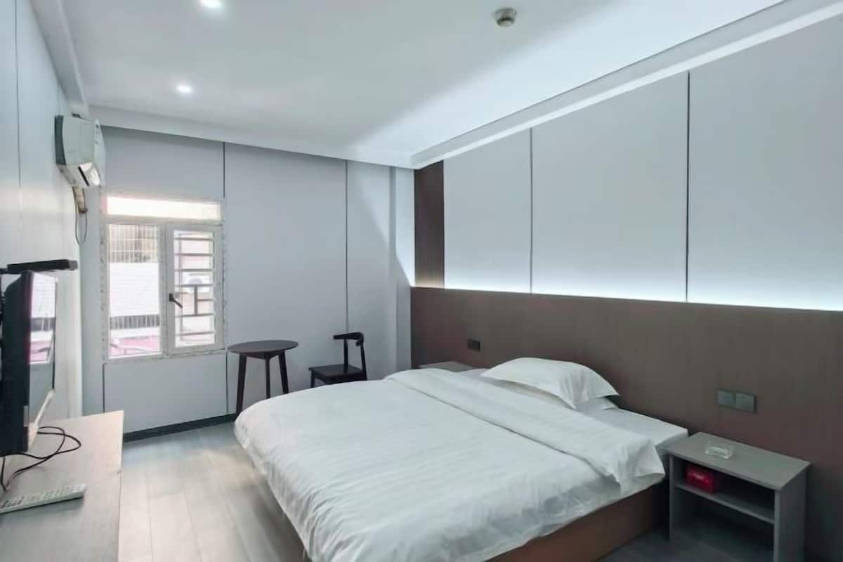 A modern hotel room with a large bed, a small desk, a chair, and a window with blinds.