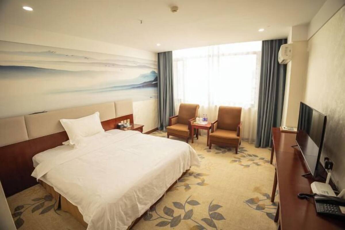 Une chambre d’hôtel comprenant un lit, deux fauteuils, un bureau avec un ordinateur et une grande fenêtre avec des rideaux.