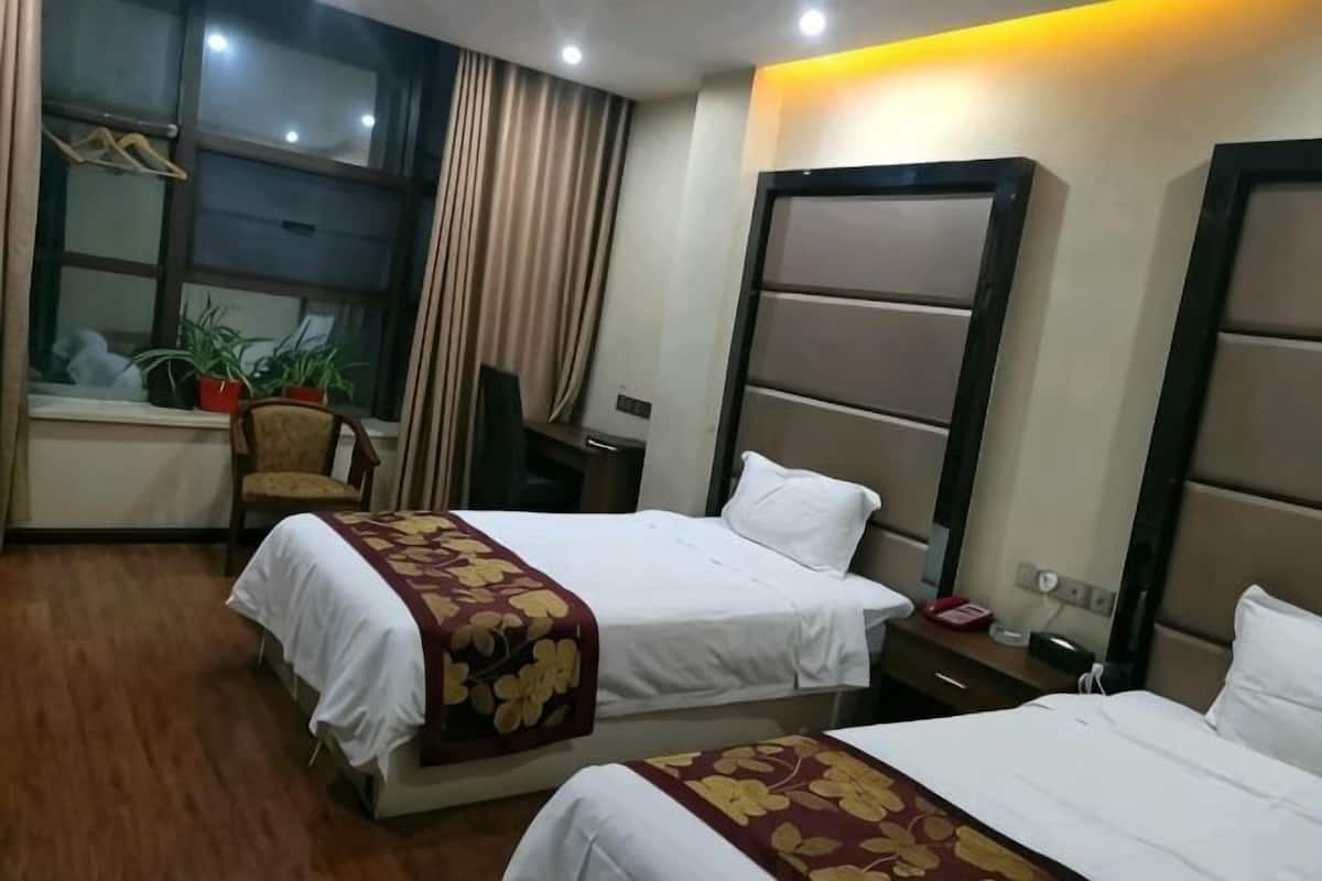Habitación de hotel con dos camas, un escritorio, una silla, un ventanal con cortinas y una planta en maceta.