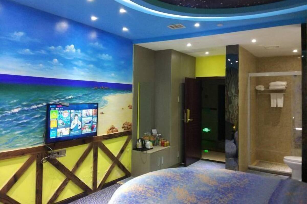 Habitación de hotel con una televisión grande en la pared, una cama con colcha estampada en azul y blanco, y un baño con ducha e inodoro.