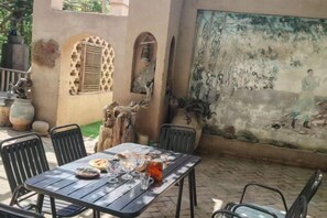 An outdoor dining area with a table set for two, surrounded by chairs, a mural on the wall, and a decorative tree trunk. - Anargvl Manor (Turpan)