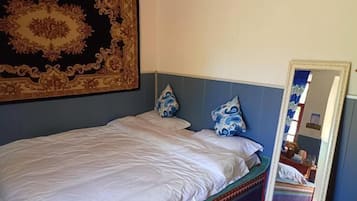 Een bed met wit beddengoed, twee gestreepte kussens, een groot versierd wandtapijt en een spiegel die een kamer met een gestreept vloerkleed reflecteert.