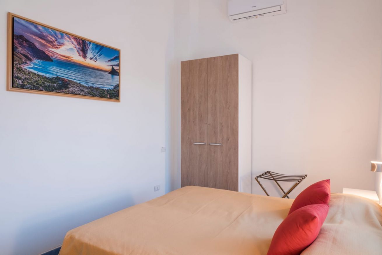A bedroom with a bed, a wooden wardrobe, a chair, and a painting of a coastal landscape.