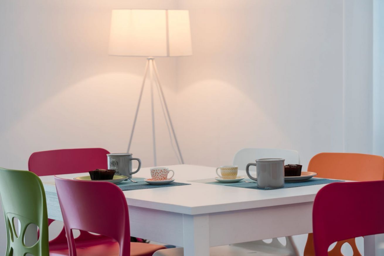 Un comedor con una mesa blanca, sillas coloridas y una lámpara de pie.