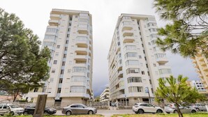 Un immeuble résidentiel de grande hauteur, doté de nombreux balcons, d’un parking avec des voitures et entouré de verdure.
