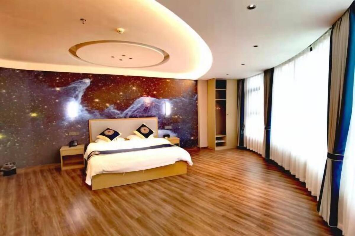 Un dormitorio moderno con techo curvo, una cama con un mural de pared con tema galáctico y ventanas amplias con cortinas.