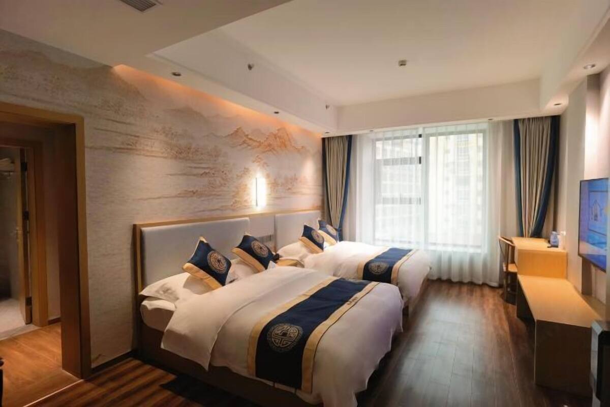 Una habitación de hotel con dos camas, suelo de madera, un ventanal grande con cortinas y un televisor montado en la pared.