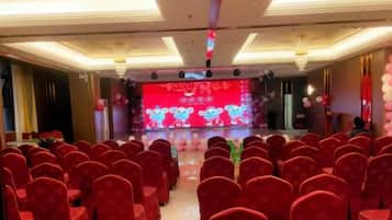 A banquet hall with rows of tables and chairs, a stage with a red backdrop, and a chandelier.