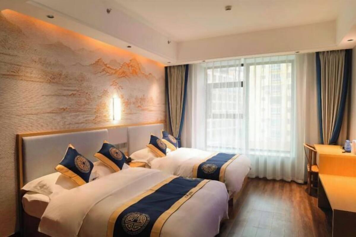 Habitación de hotel con dos camas, un ventanal con cortinas, un mural en la pared y una mesita.