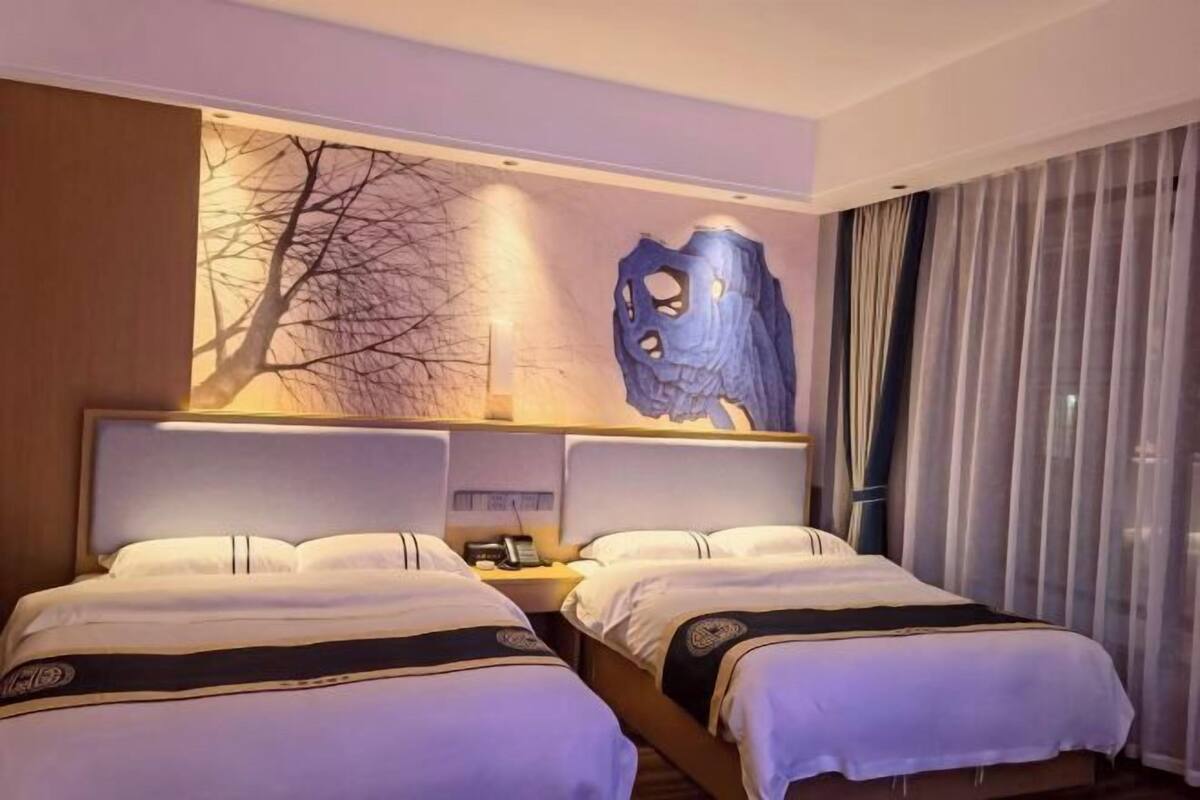 Una habitación de hotel con dos camas, una mesita de noche, una lámpara de pared y un ventanal con cortinas.