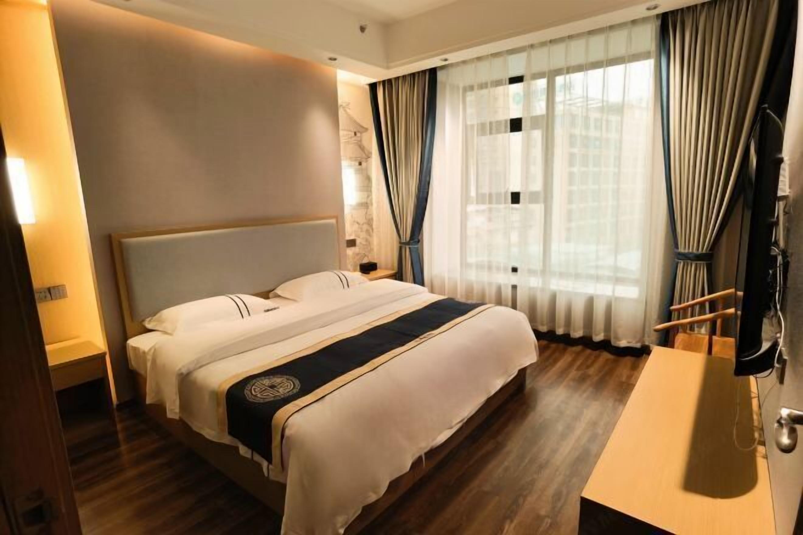 Habitación de hotel con una cama grande, piso de madera, mesita de noche, televisor y ventana con cortinas.