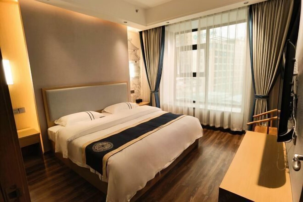 A hotel room with a large bed, a wooden floor, a bedside table, a TV, and a window with curtains.