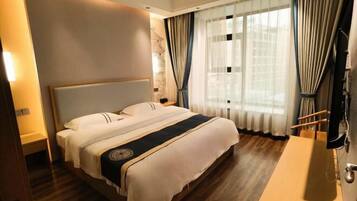 Habitación de hotel con una cama grande, piso de madera, mesita de noche, televisor y ventana con cortinas.