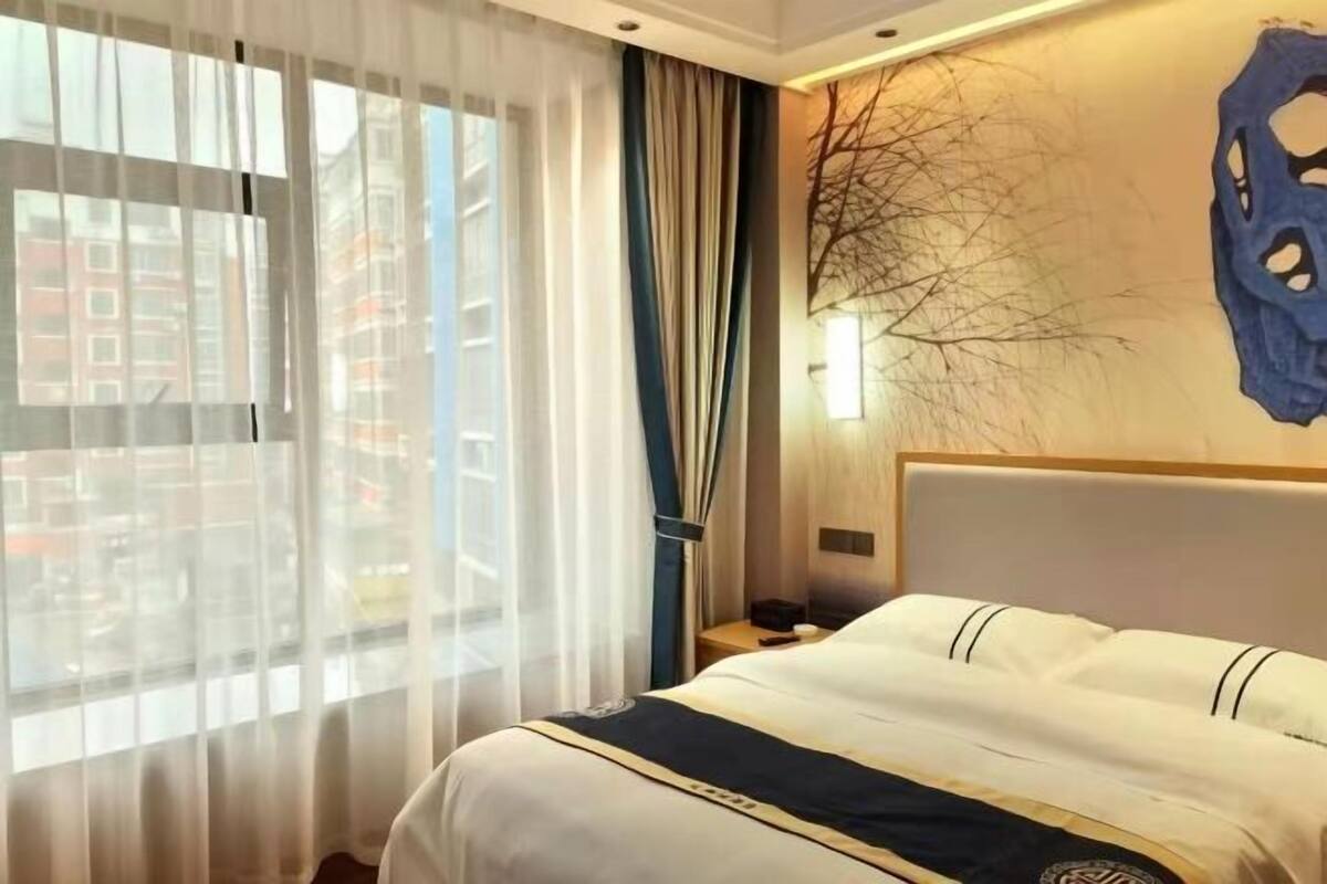 Una habitación de hotel con una cama, una mesita de noche, una lámpara y una ventana con cortinas translúcidas.