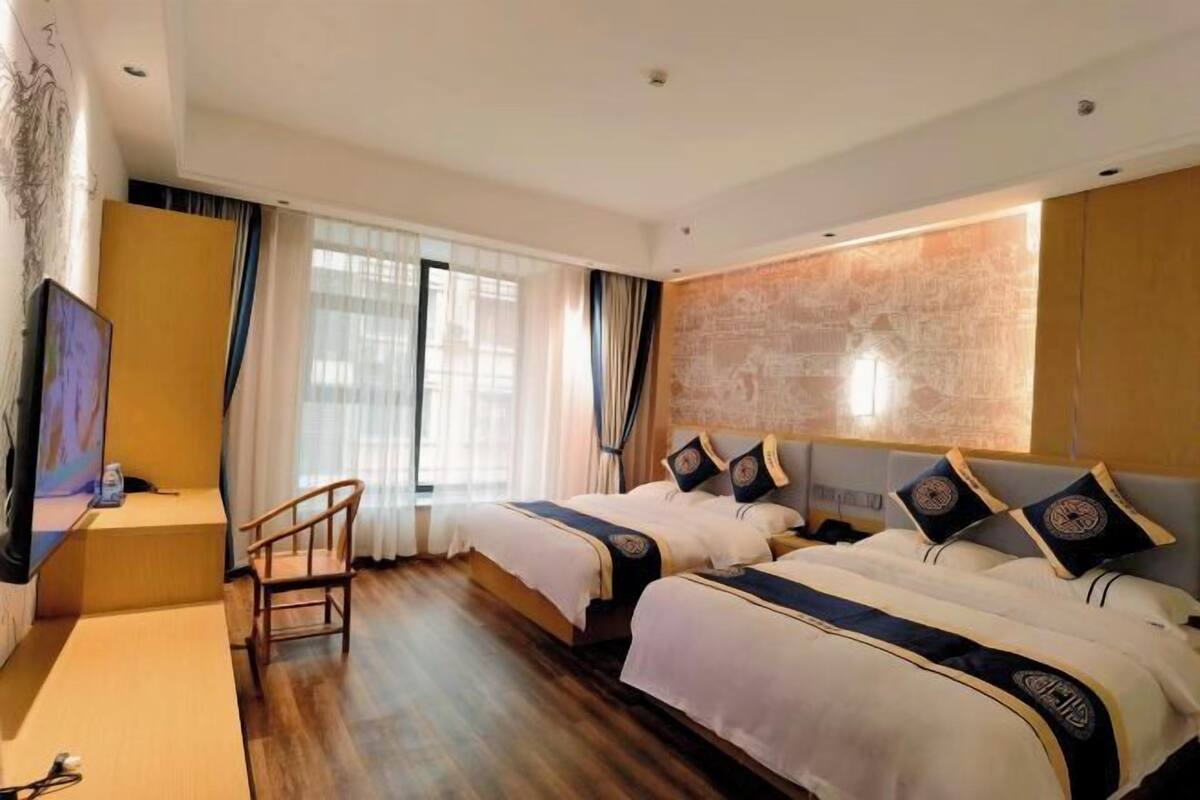 Una habitación de hotel con dos camas, televisión, una silla de madera y un ventanal con cortinas.