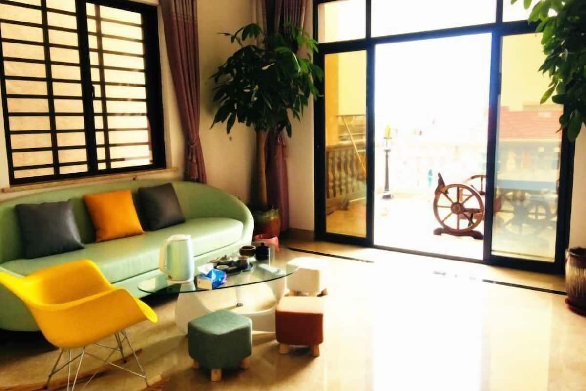 A living room with a green sofa, yellow chair, and small ottomans. There is a large window with a view of a building and a ship's wheel.
