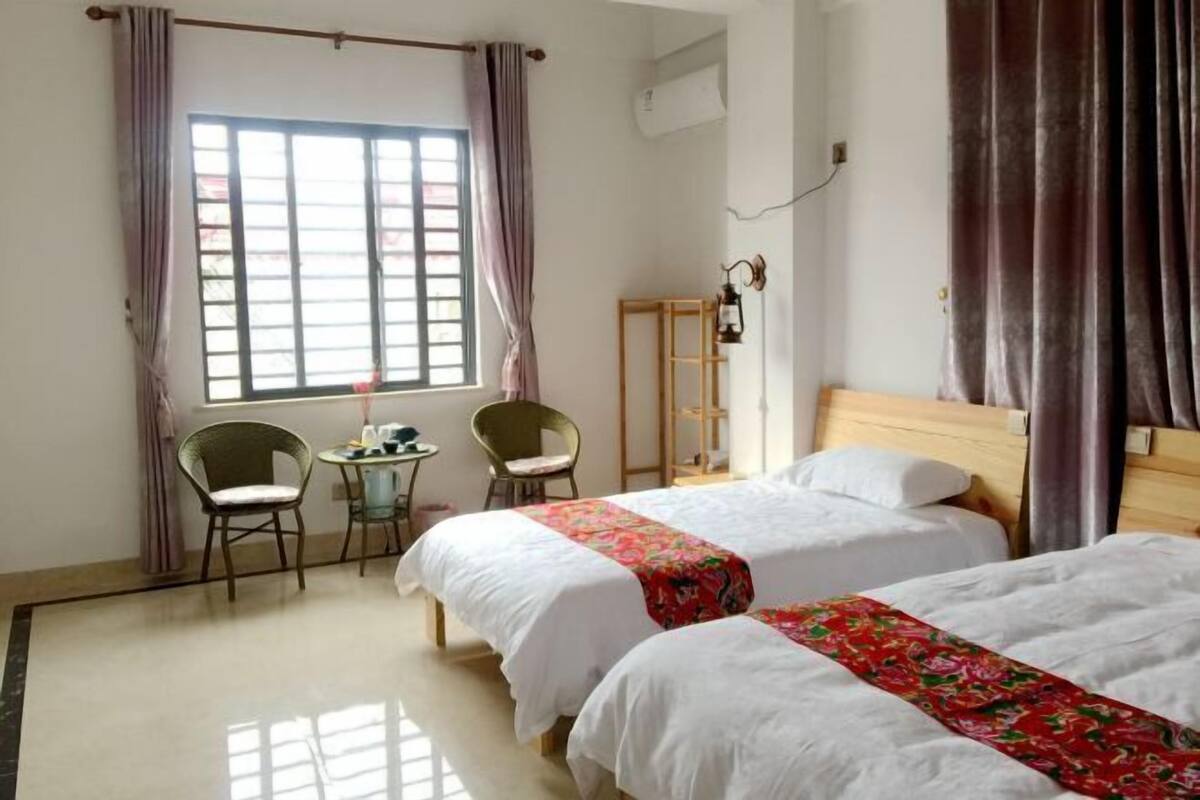 A hotel room with two beds, a small table with chairs, a lamp, and a window with curtains.