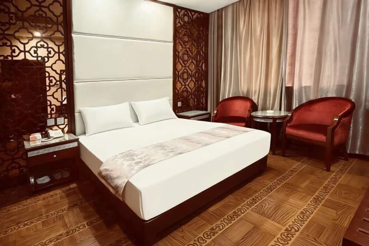 A hotel room with a large bed, two red chairs, a small table, a nightstand, and patterned curtains.