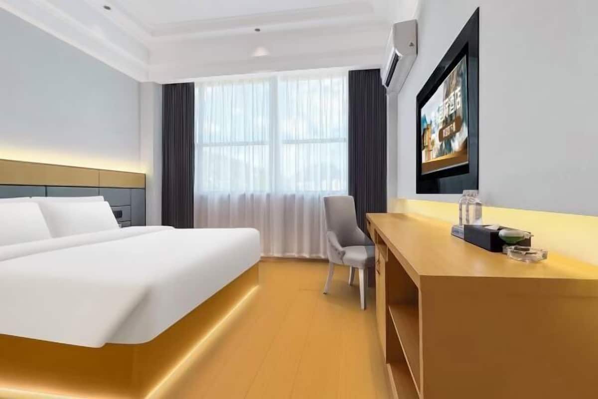 Quarto de hotel com cama, escrivaninha, cadeira e uma janela grande com cortinas.