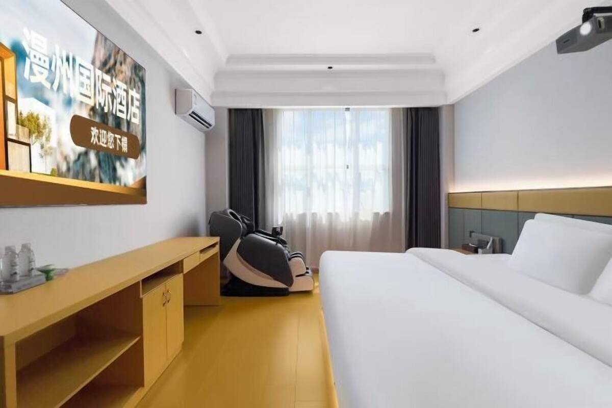 Quarto de hotel com cama, cadeira de massagem, escrivaninha e uma grande janela com cortinas.