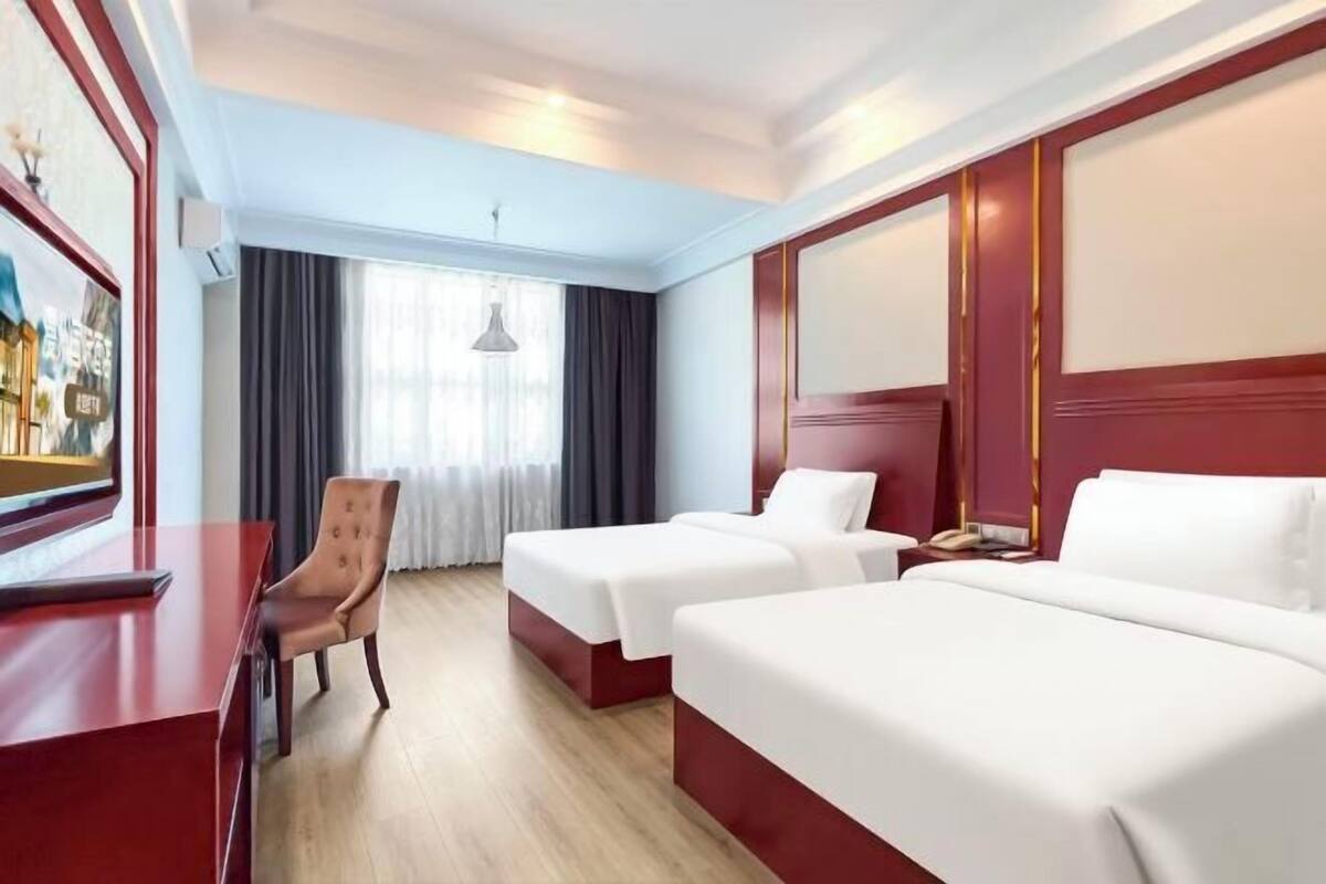 Quarto de hotel com duas camas, uma escrivaninha, uma cadeira e uma janela grande com cortinas.