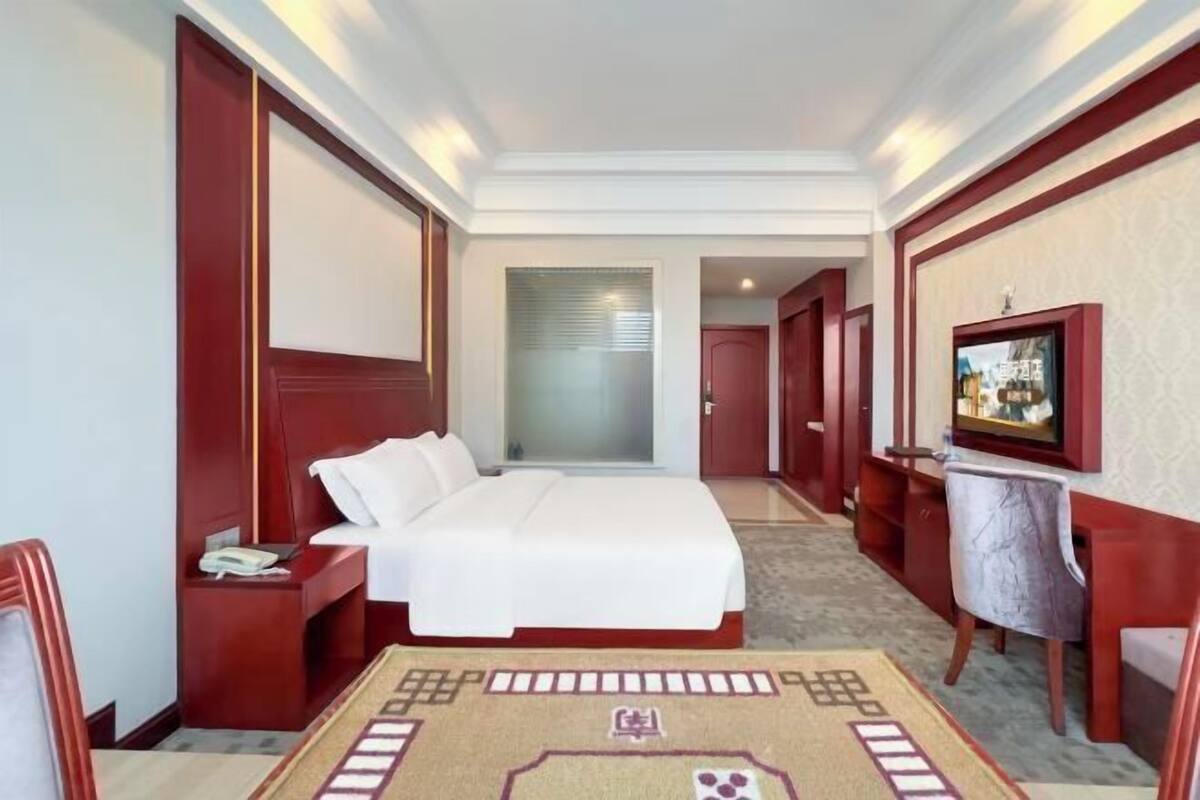 Quarto de hotel com uma cama grande, uma mesa com cadeira, televisão e um tapete com padrão geométrico.