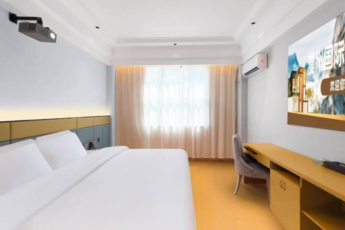 Quarto de hotel com cama, escrivaninha, cadeira, projetor e uma grande janela com cortinas.