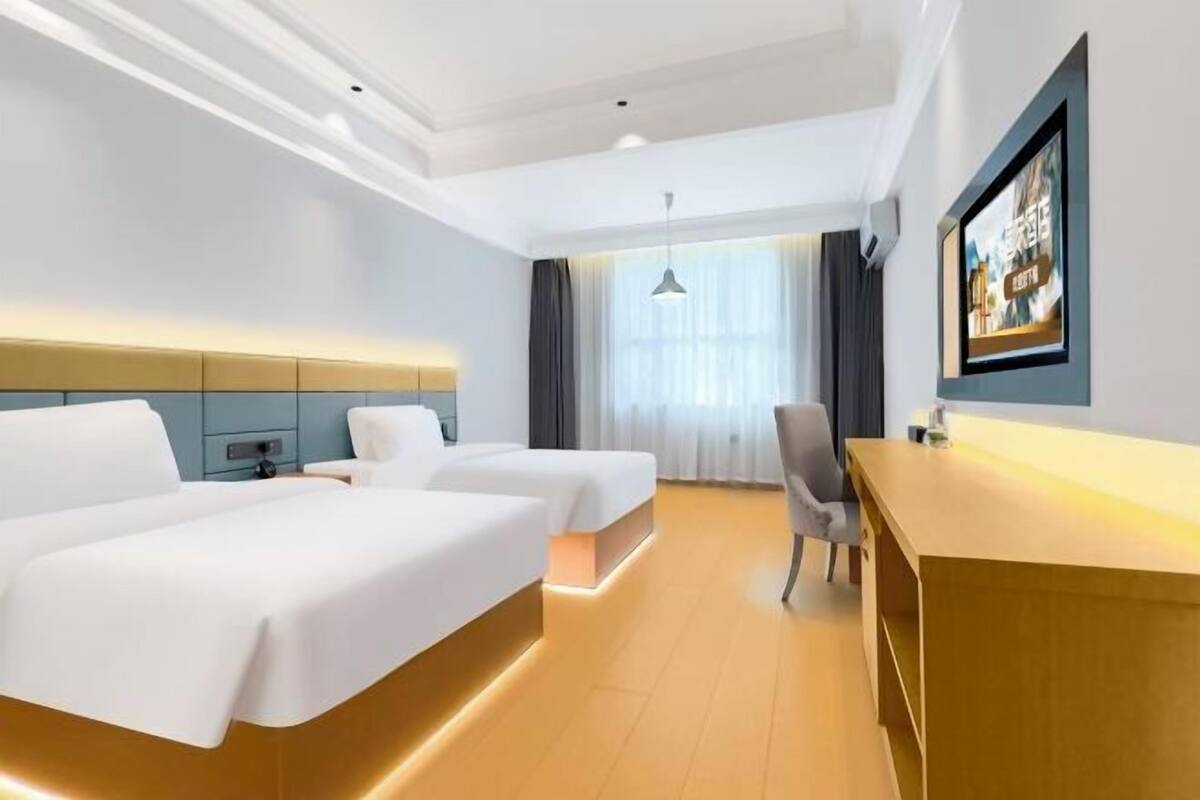 Quarto de hotel com duas camas, uma mesa, uma cadeira, uma janela grande com cortinas e um quadro na parede.