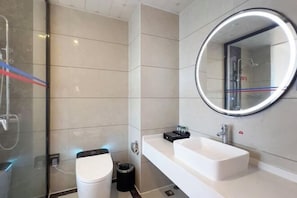 Un baño moderno con un lavamanos blanco, un espejo redondo con luz y una cabina de ducha de vidrio.
