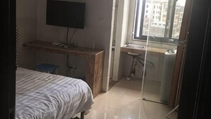Une chambre à coucher comprenant un lit, un bureau, une chaise, un téléviseur et une fenêtre donnant sur des immeubles.